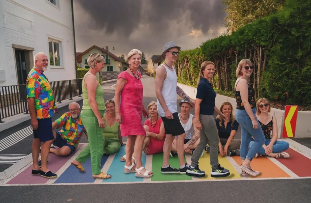 12 Personen, sechs davon sitzend und sechs davon stehend auf einen in Regenbogenfarben gefärbten Zebrastreifen