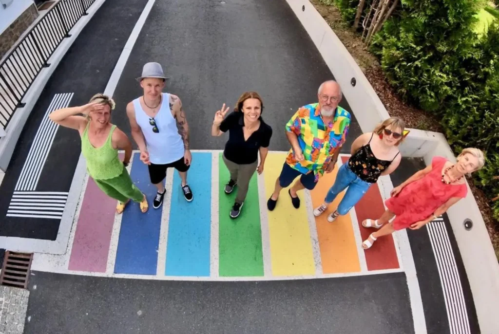 Sechs Personen zum Großteil bunt gekleidet auf einen in Regenbogenfarben gefärbten Zebrastreifen